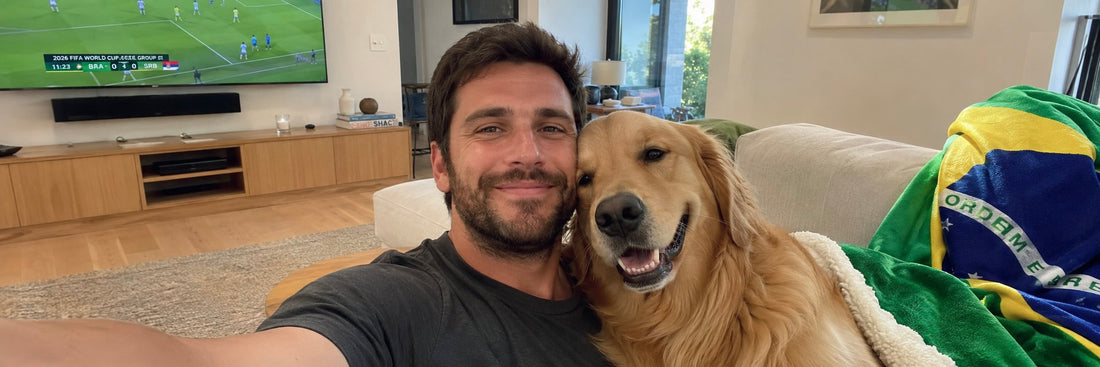 A smiling man and his Golden Retriever dog draped in a soccer jersey watching a match on TV at home.