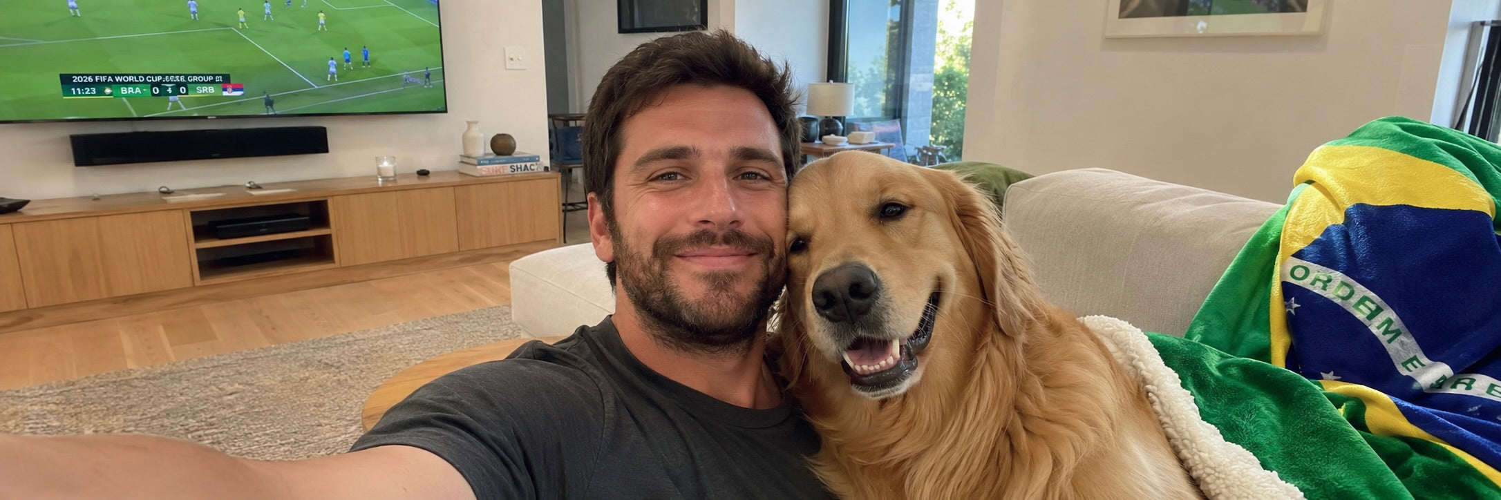 A smiling man and his Golden Retriever dog draped in a soccer jersey watching a match on TV at home.