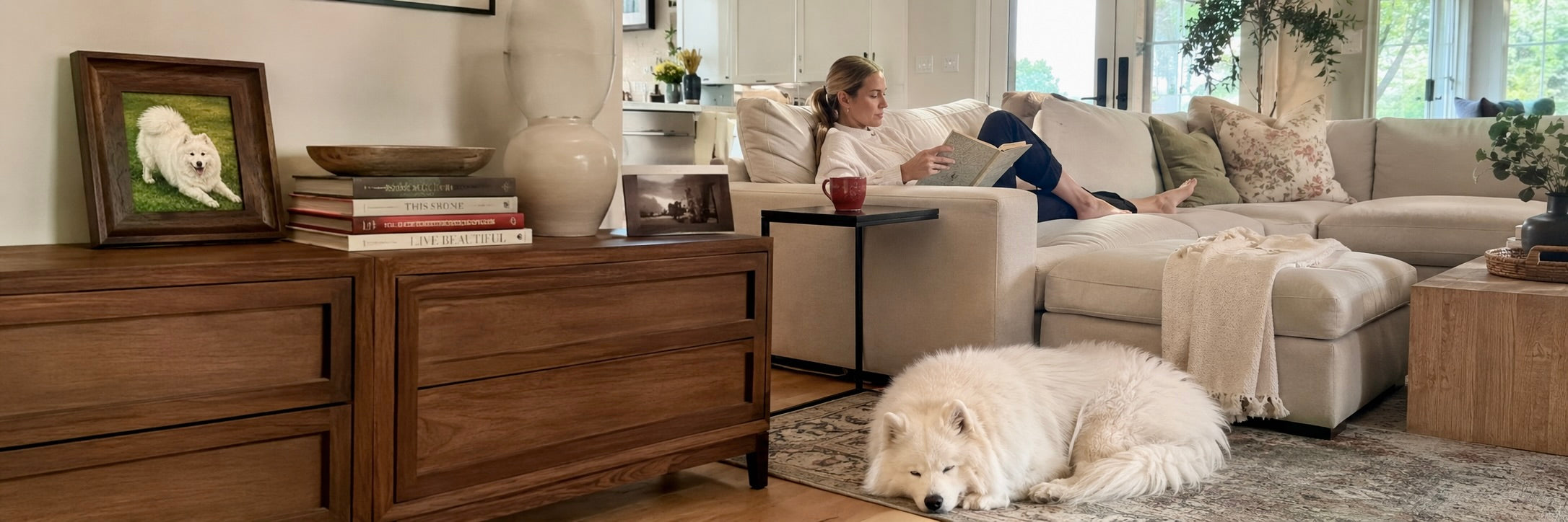 A woman sits on a couch with a white dog in a sunlit living room.
