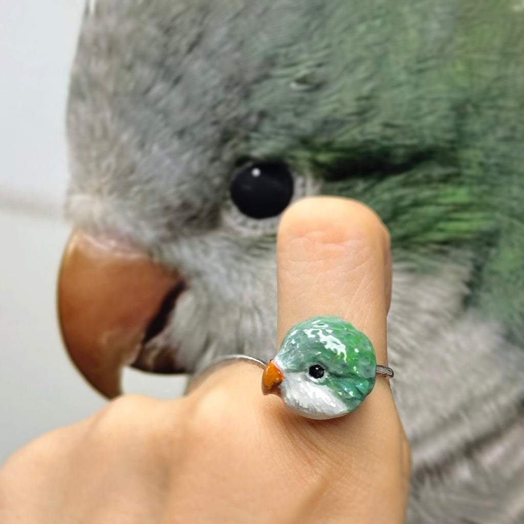 A real green parakeet next to a bespoke 3D pet portrait ring, custom hand-sculpted to perfectly replicate the bird as a luxury exotic pet jewelry keepsake.