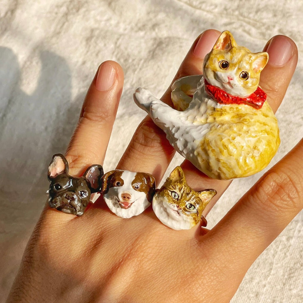 A hand showcasing multiple bespoke 3D pet portrait rings, featuring custom hand-sculpted cats and dogs as premium sentimental wedding keepsakes.