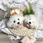 Two fluffy white dogs in a woven basket with flowers and a gift on a textured blanket.