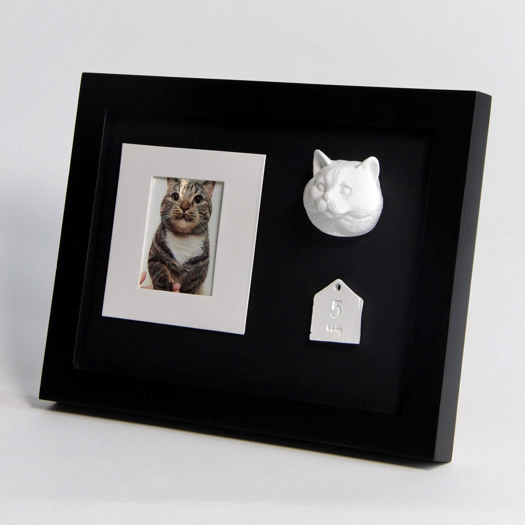 Black framed display with a photo of a cat, a white cat figurine, and a tag inside.