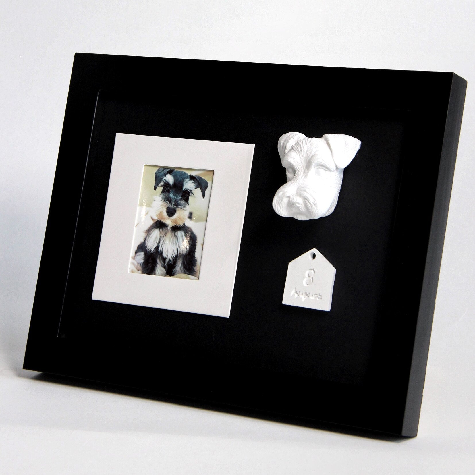 Black frame with a photo of a dog, a white dog sculpture, and a tag inside.