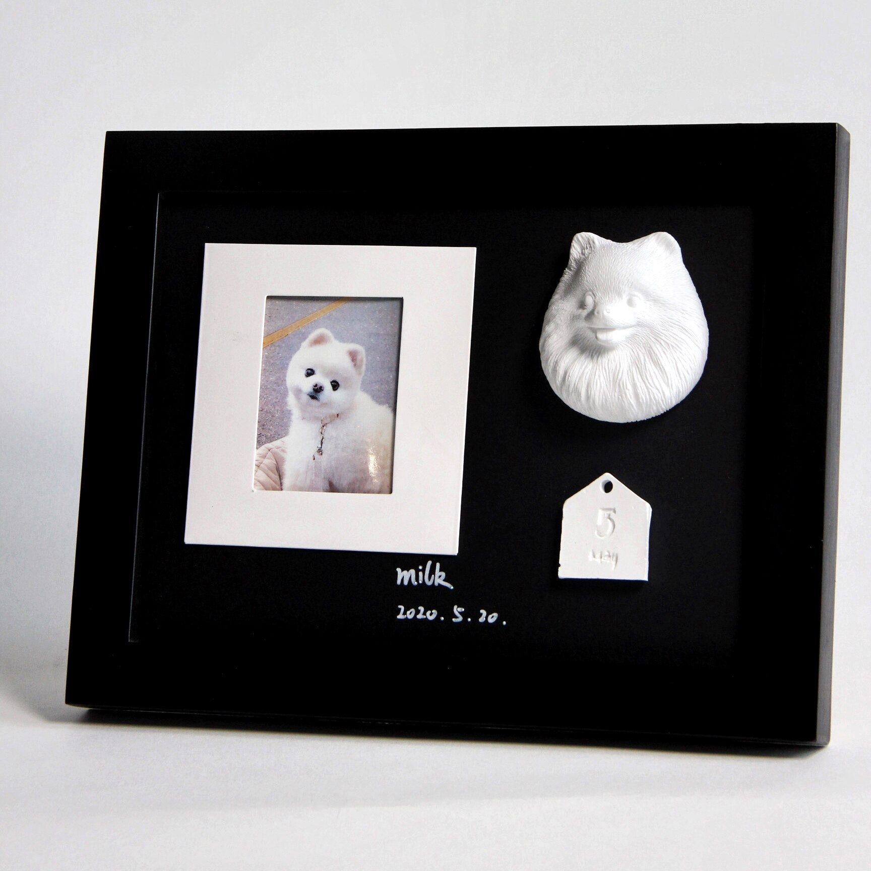 Black frame with a photo of a dog, a white ceramic tag, and a white ceramic animal sculpture on a light gray background.