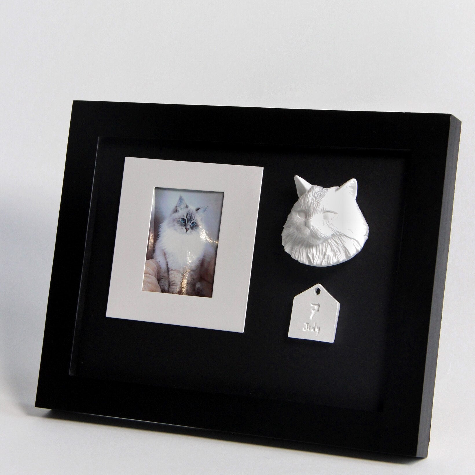 Black framed display with a photo of a cat, a 3D cat sculpture, and a cat-shaped tag.