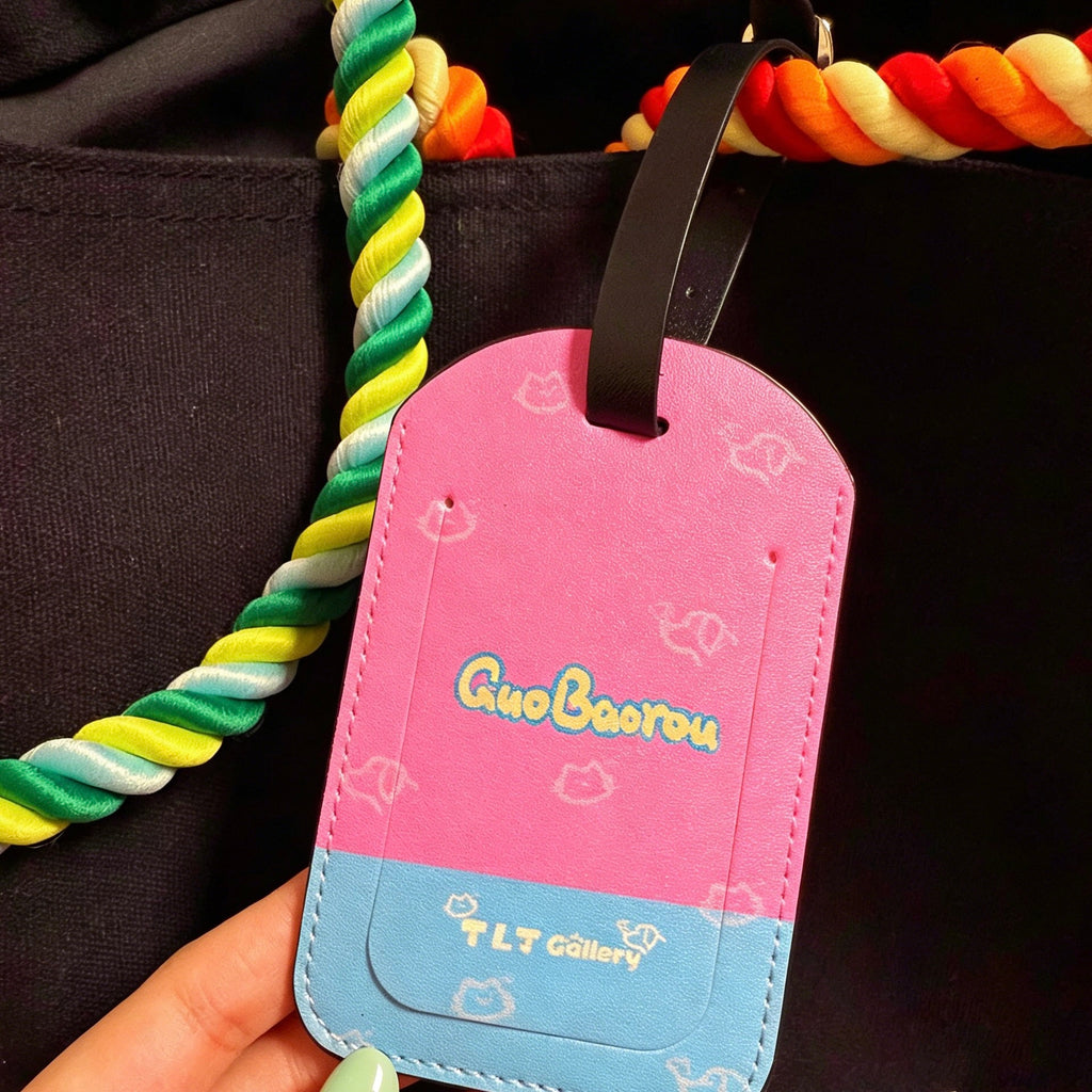 Pink and blue luggage tag with 'GiuBoerou' branding held by a hand against a dark background.