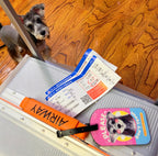Small dog standing next to an open suitcase with travel documents and a luggage tag on a wooden floor.