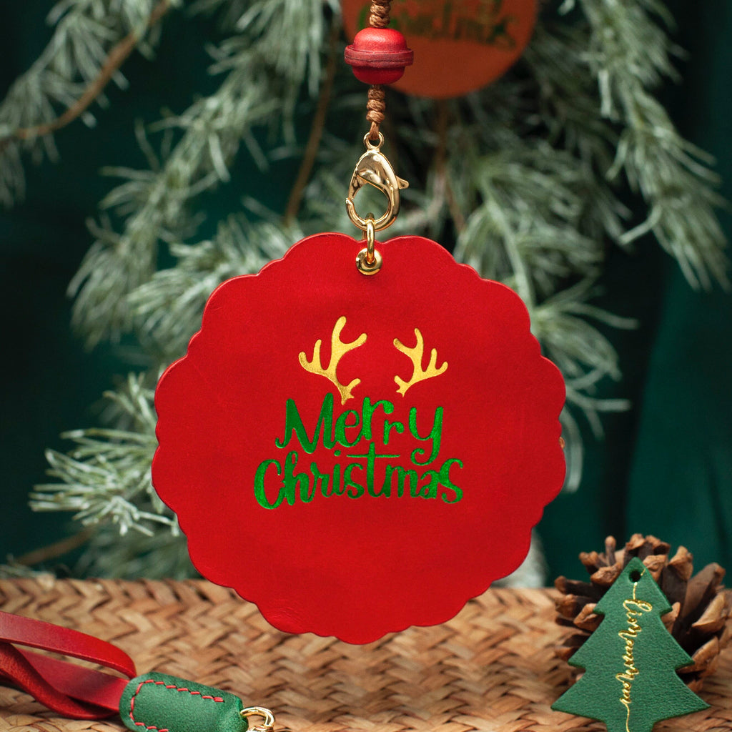 Red Christmas ornament with 'Merry Christmas' text and antlers, hanging against a green foliage background.