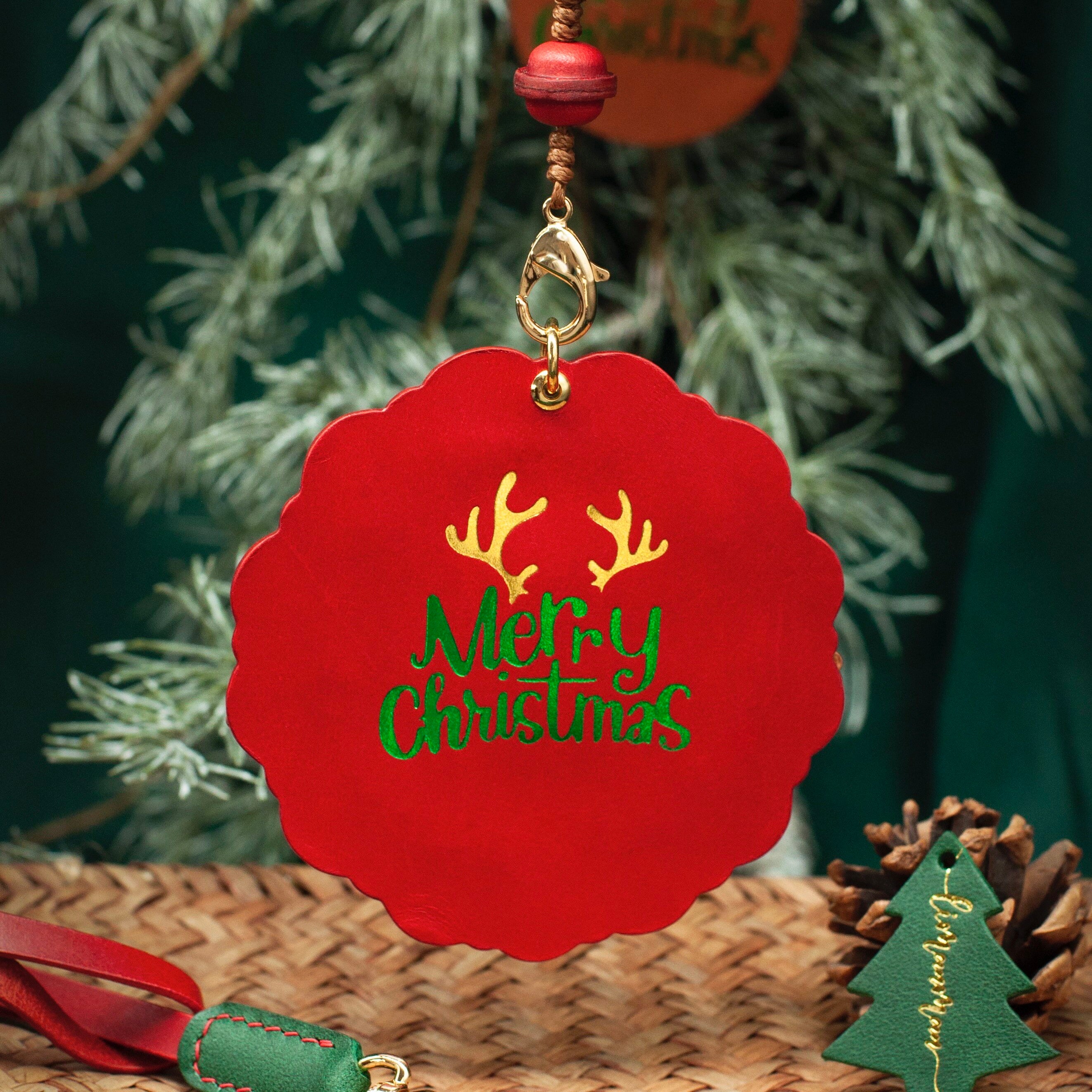Red Christmas ornament with 'Merry Christmas' text and antlers, hanging against a green foliage background.