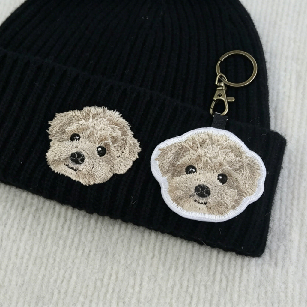 Black knit beanie with embroidered dog faces and a matching keychain on a light gray background.