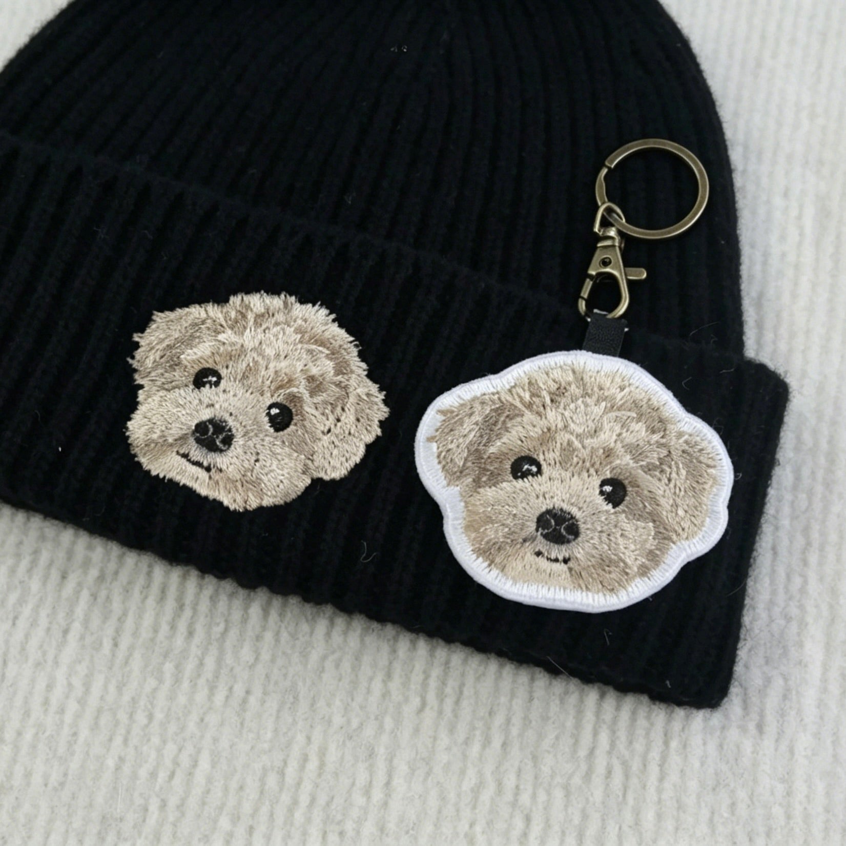 Black knit beanie with embroidered dog faces and a matching keychain on a light gray background.