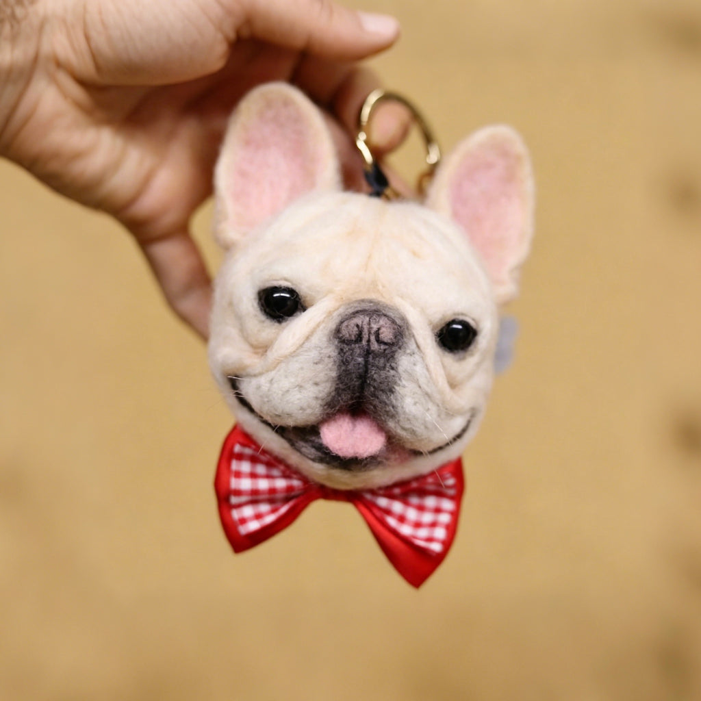 Felted animal keychain of a dog with a red bow tie held by a hand against a beige background