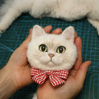 Hand holding a small white cat-shaped plush toy with a red checkered bow on a blue cutting mat.