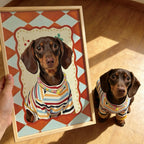 Dachshund wearing a striped outfit next to a framed painting of itself