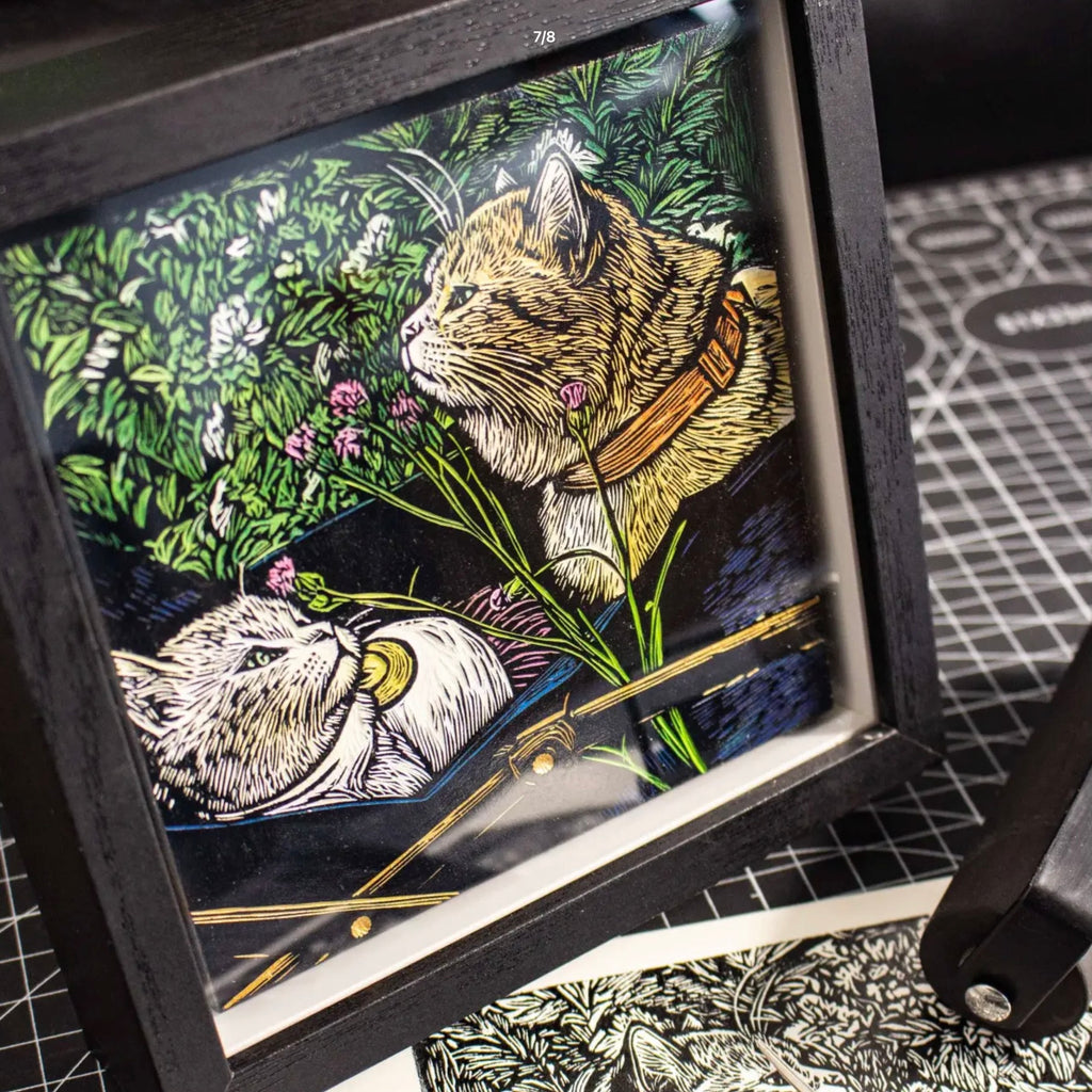 Framed artwork of two cats in a garden setting with flowers and leaves.