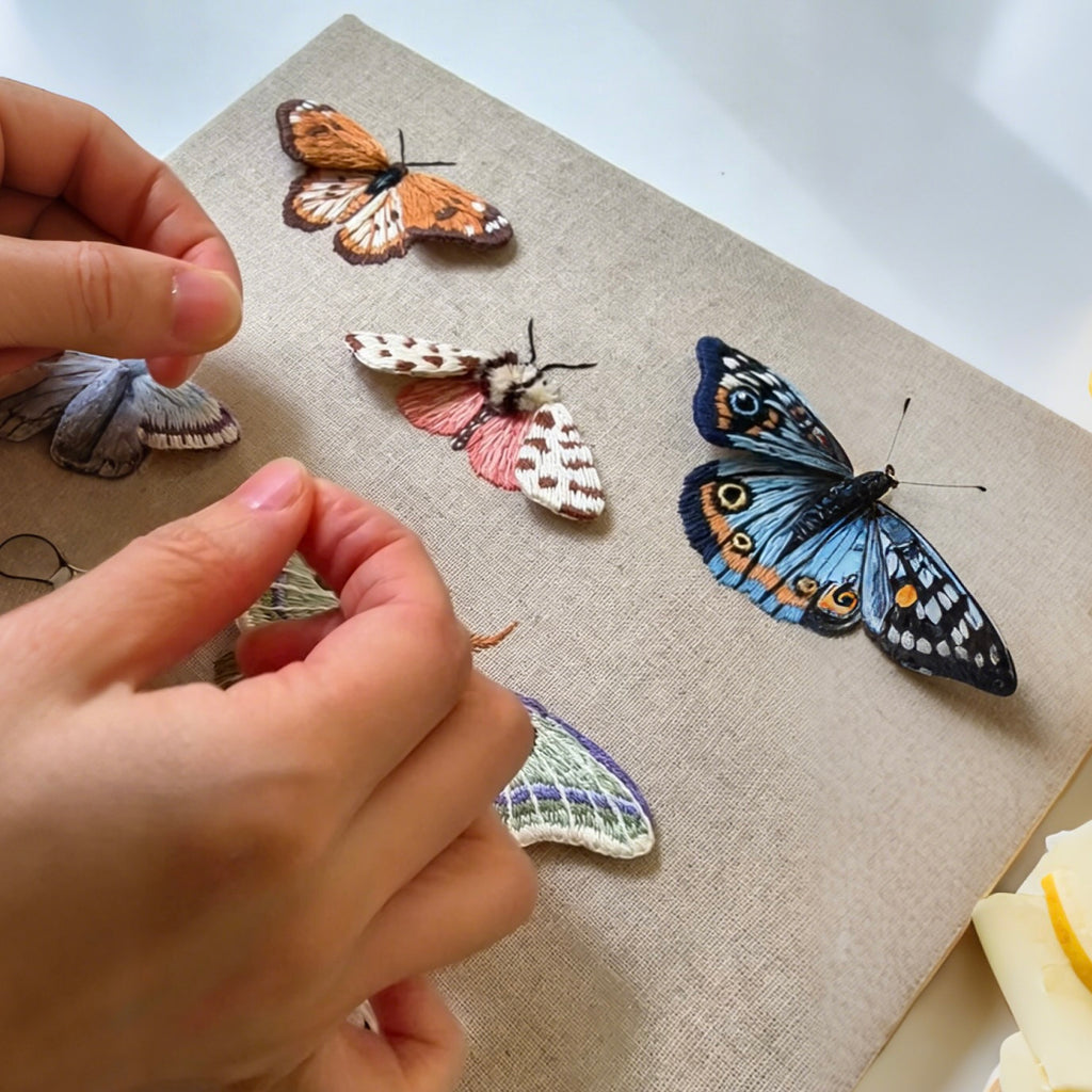 Hand embroidery process showing realistic thread texture of a custom butterfly brooch, being delicately adjusted with a traditional needle tool.