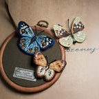 A luxury hand-embroidered butterfly art display featuring a vibrant blue butterfly and an orange butterfly framed in a wooden hoop with a vintage specimen label, showcasing realistic textile craftsmanship.
