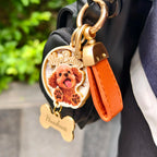 A close-up shot of a custom 3D wooden pet portrait keychain styled as a bag charm, featuring a realistic 3D relief of a brown poodle with the text "My Baby". This personalized memorial keepsake includes a vibrant orange leather strap, gold hardware, and a wooden bone tag engraved with the pet's name "Huahua".