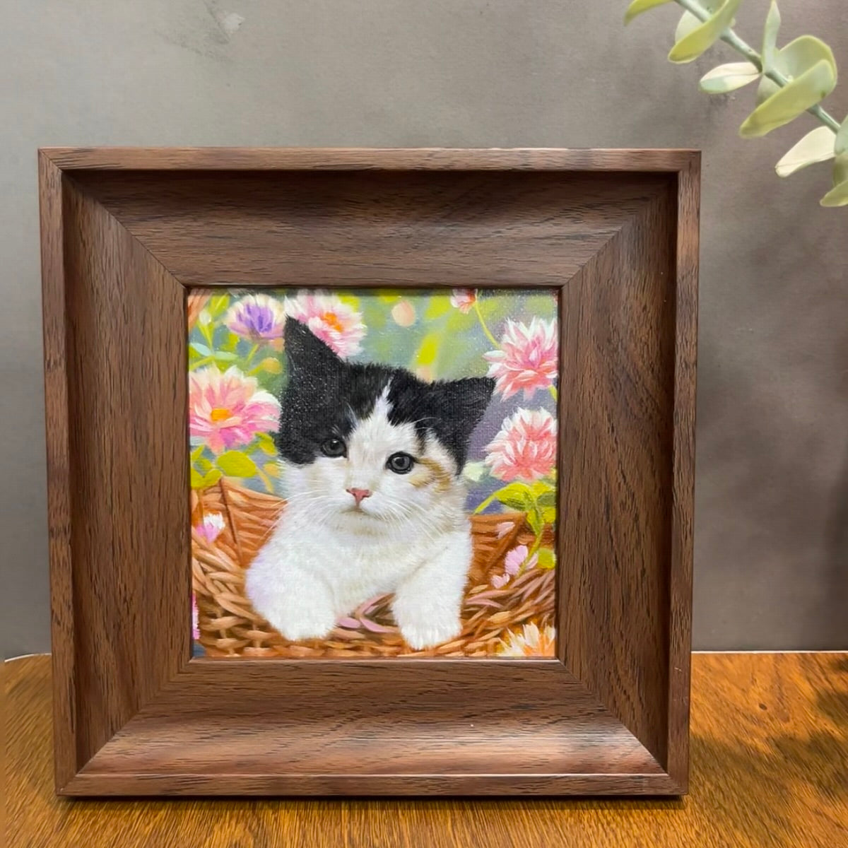 Framed artwork of a cat in a basket with flowers on a wooden surface