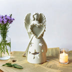 White angel figurine with a heart, placed on a table with a candle and flowers.