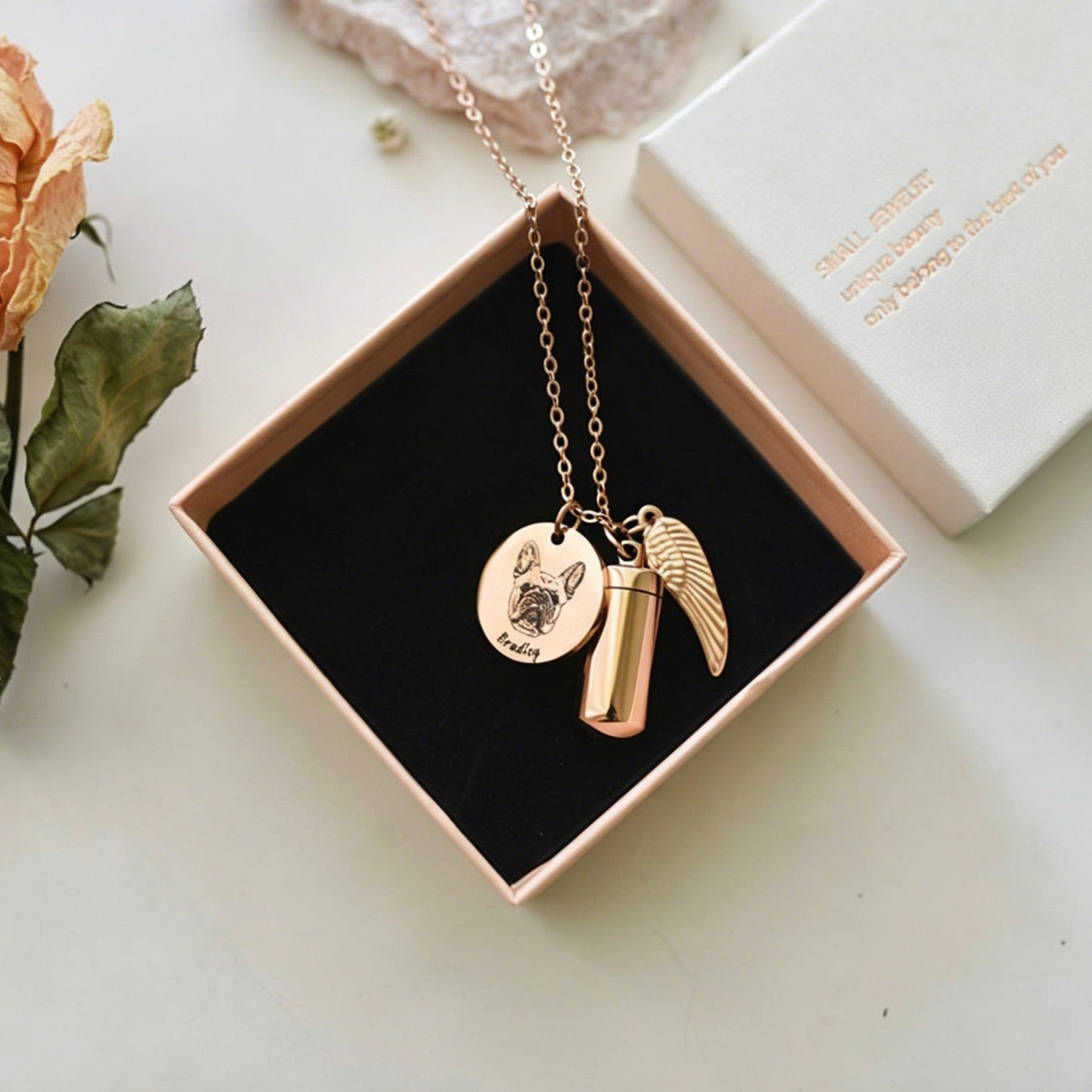 Gold necklace with pendant and wings in a box on a light surface with a rose and card.