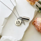 Silver necklace with a pendant and cylindrical charm on a white surface with dried flowers.