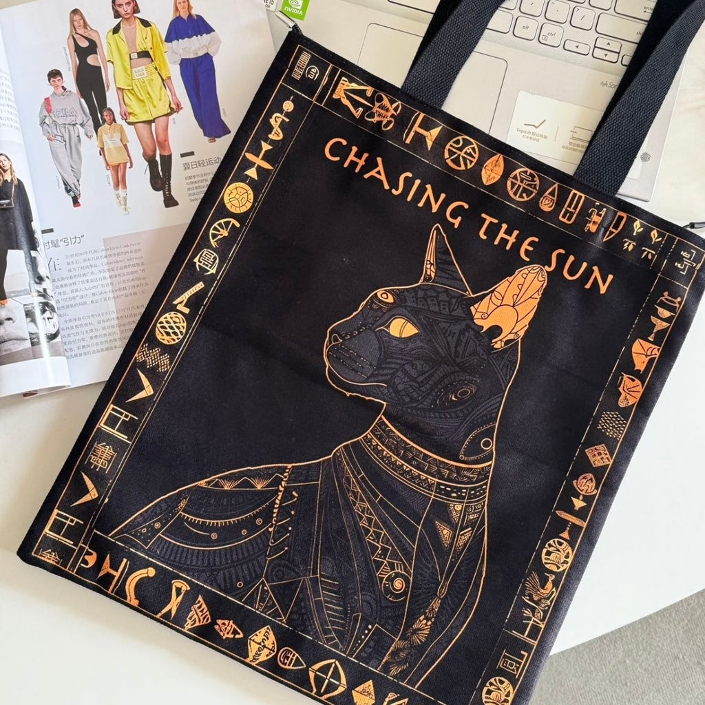 Black tote bag with gold cat design and 'Chasing the Sun' text on a desk.