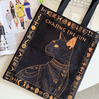 Black tote bag with gold cat design and 'Chasing the Sun' text on a desk.