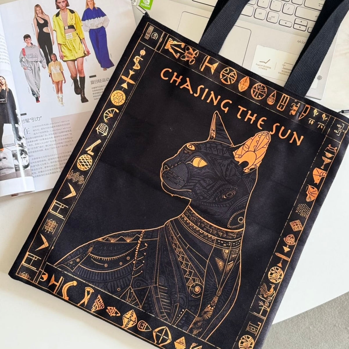 Black tote bag with gold cat design and 'Chasing the Sun' text on a desk.