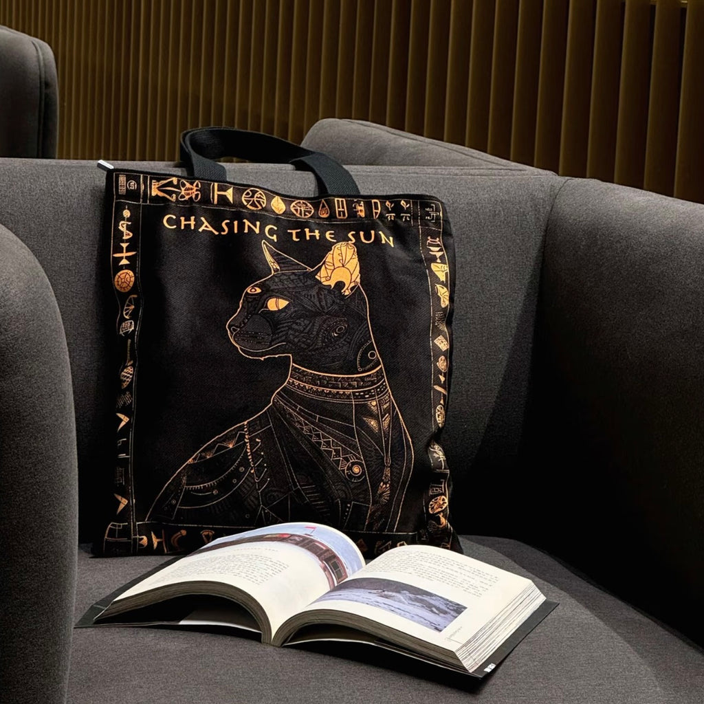 Tote bag with cat design and 'Chasing the Sun' text on a gray couch next to an open book.
