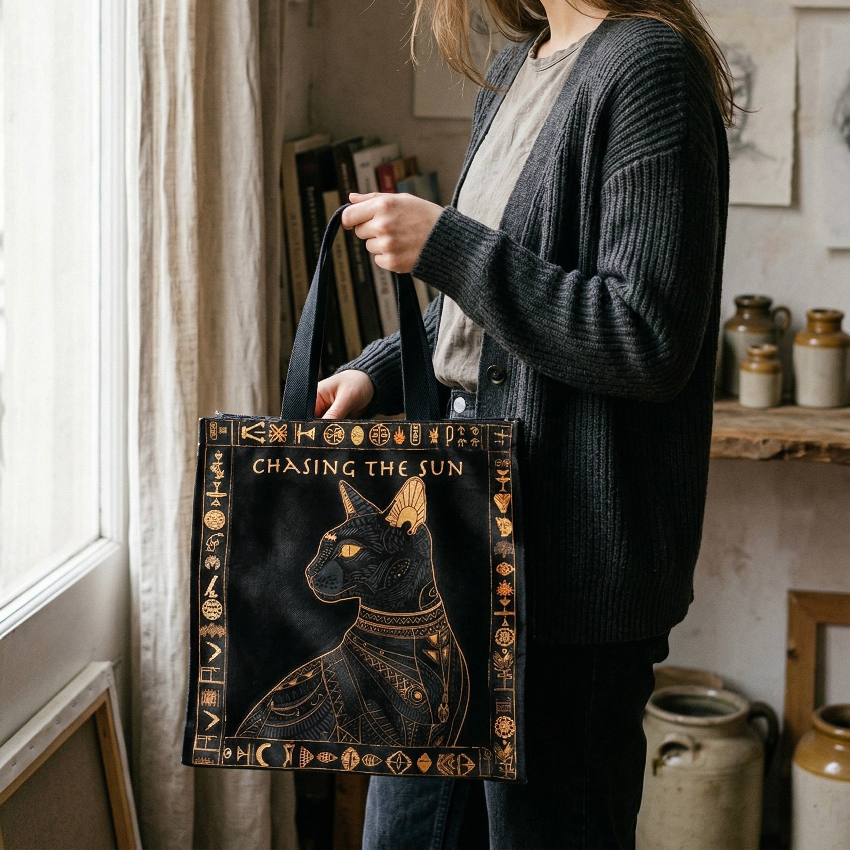 Person holding a tote bag with a cat design and 'Chasing the Sun' text in a home setting.