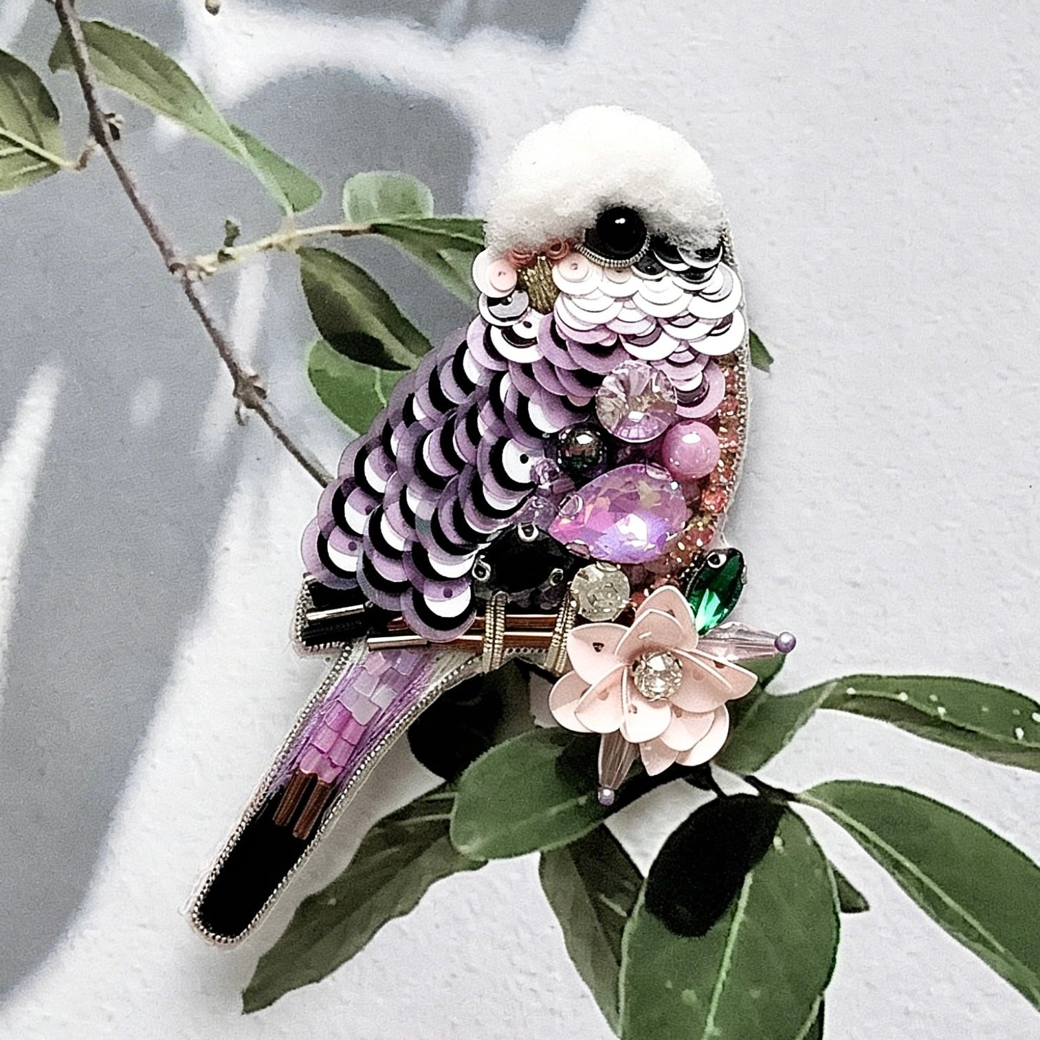 Decorative bird-shaped object with sequins and gemstones on a leafy branch.