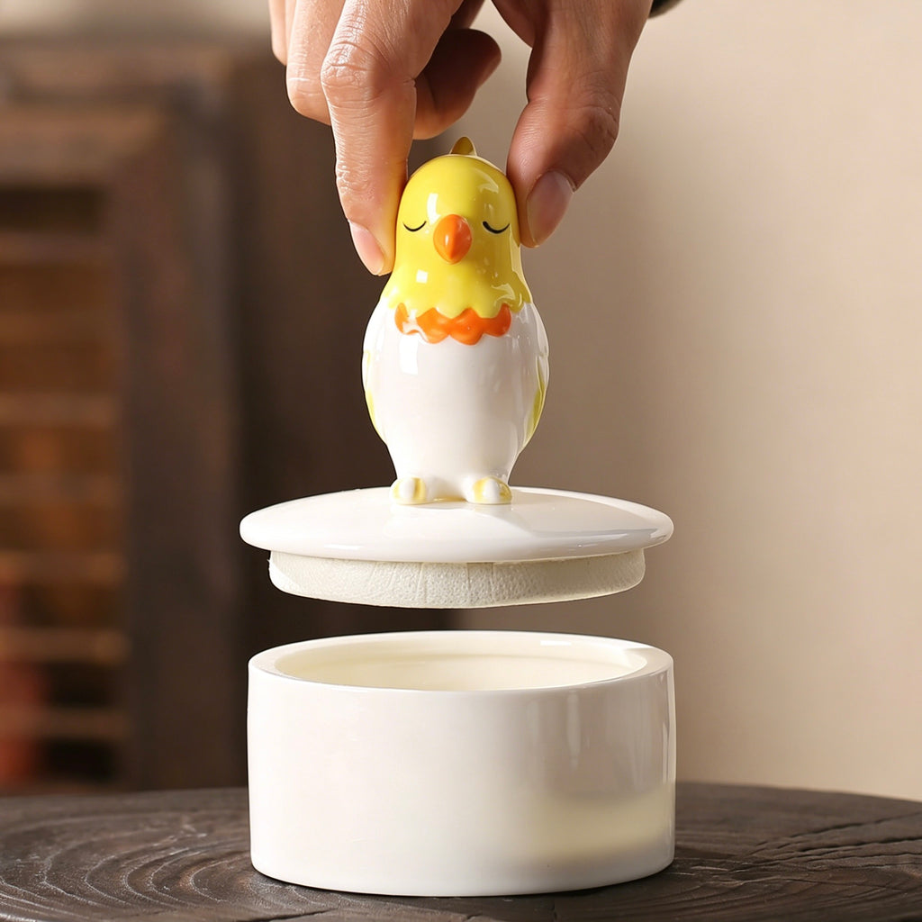 Ceramic chicken-shaped lid being lifted off a white container on a wooden surface.