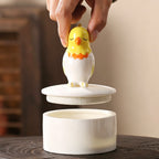 Ceramic chicken-shaped lid being lifted off a white container on a wooden surface.