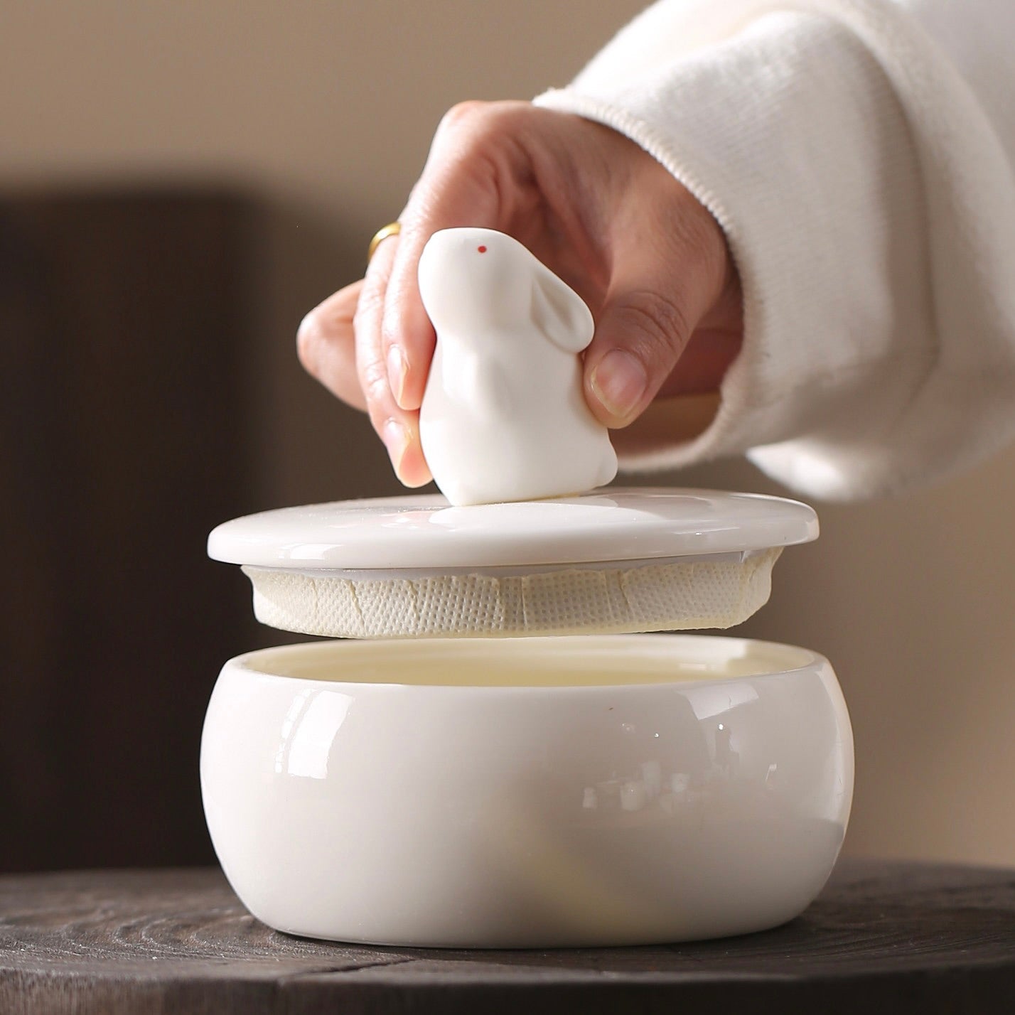 Person holding a white ceramic tea infuser with a lid on a wooden surface.