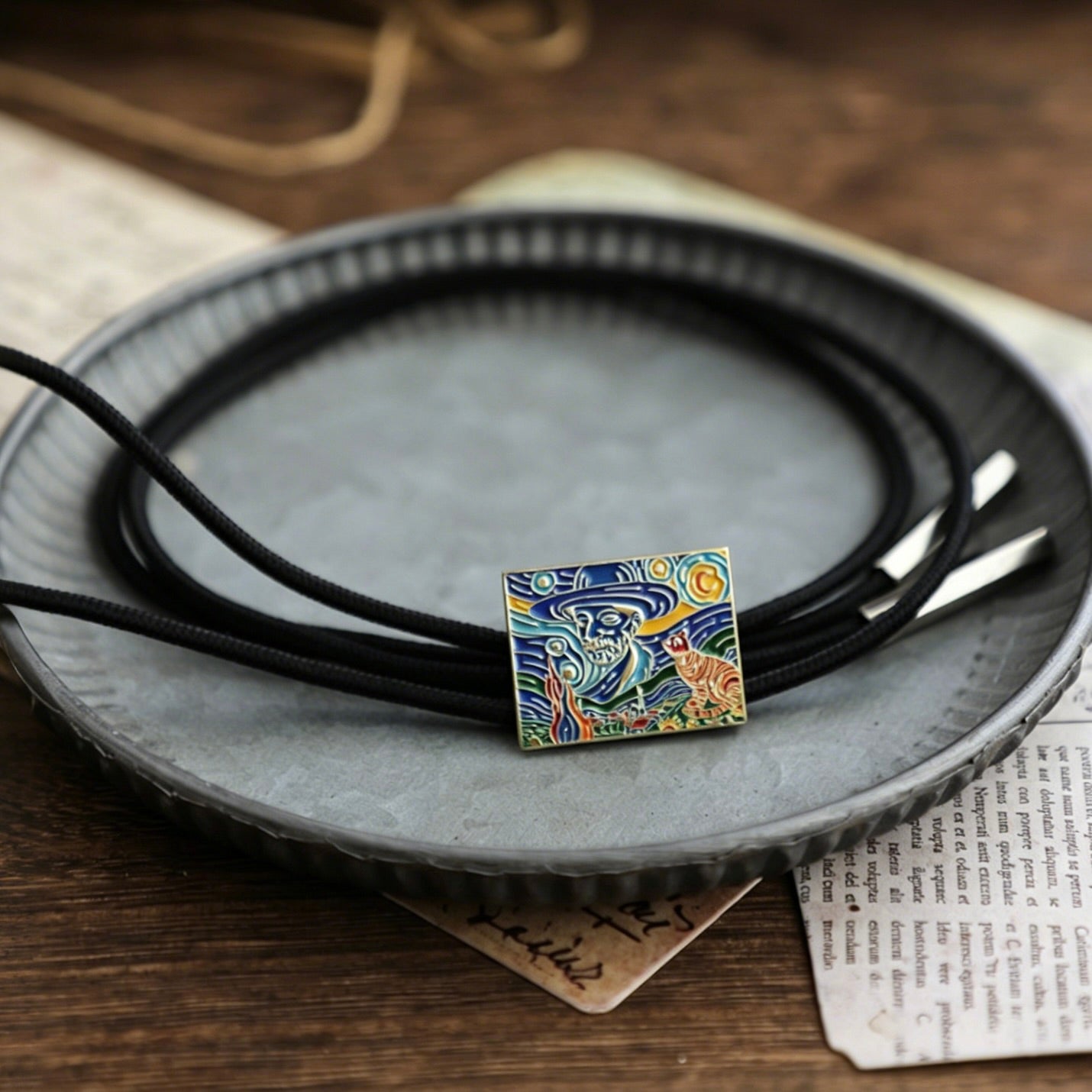 Necklace with a square pendant featuring a stylized depiction of a cat under a starry night sky, on a dark surface.