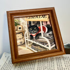 Brown framed relief portrait of a black cat in a red outfit resting on folded textiles beneath a rail