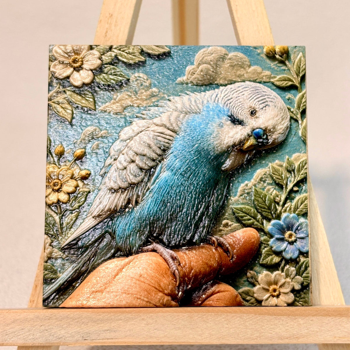 Unframed relief artwork of a blue-and-white budgie perched on a finger, surrounded by flowers and clouds