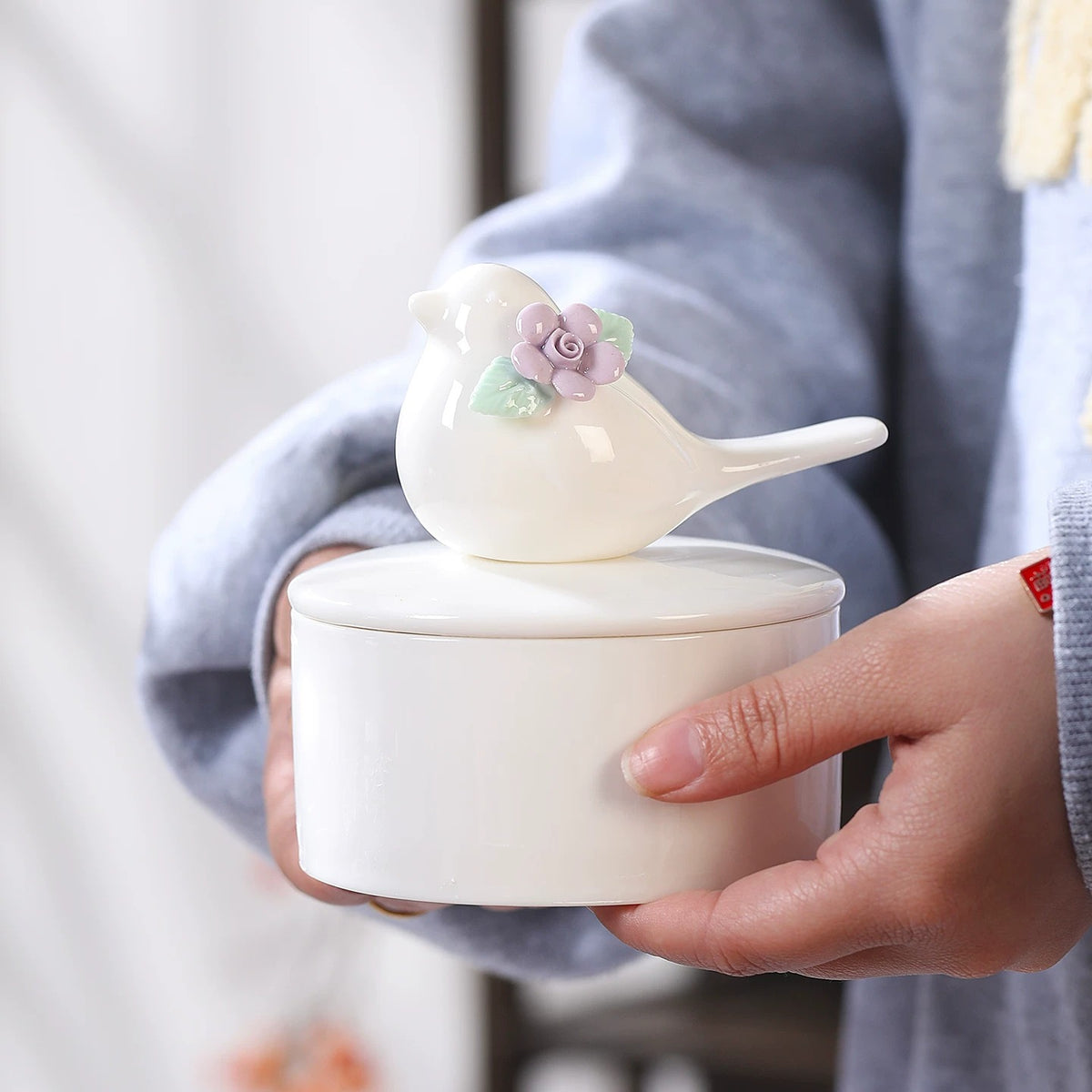 Person holding an ivory ceramic bird memorial urn with a purple flower and green leaf decoration on the lid