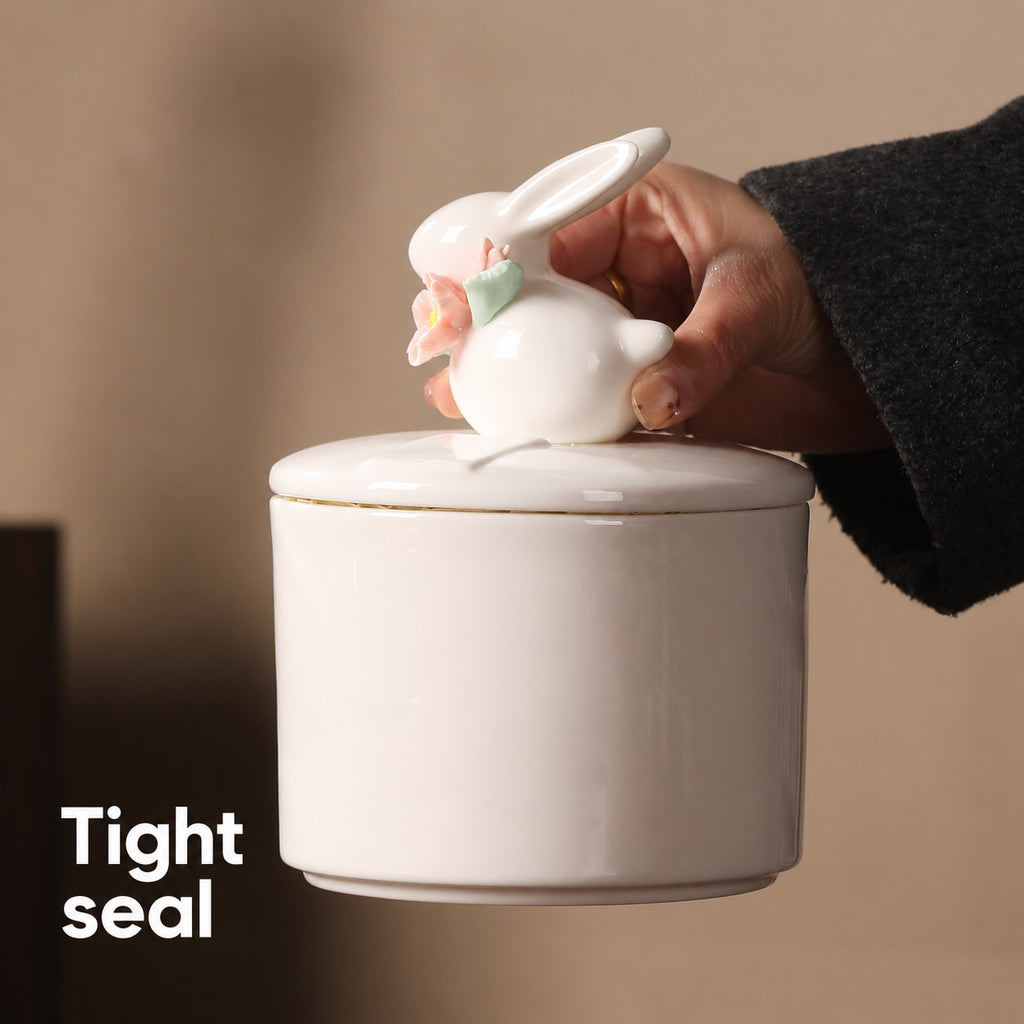 Hand holding the rabbit-shaped lid above an ivory floral ceramic rabbit memorial urn with a close-fitting lid