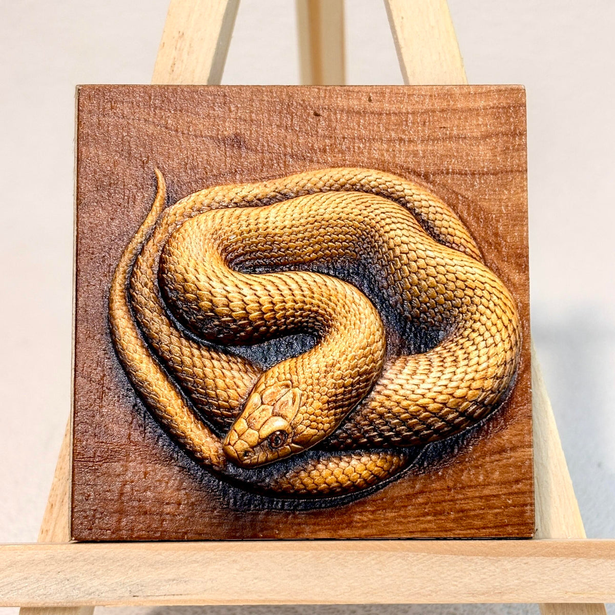 Unframed relief artwork of a coiled golden snake with raised scale details, displayed on an easel