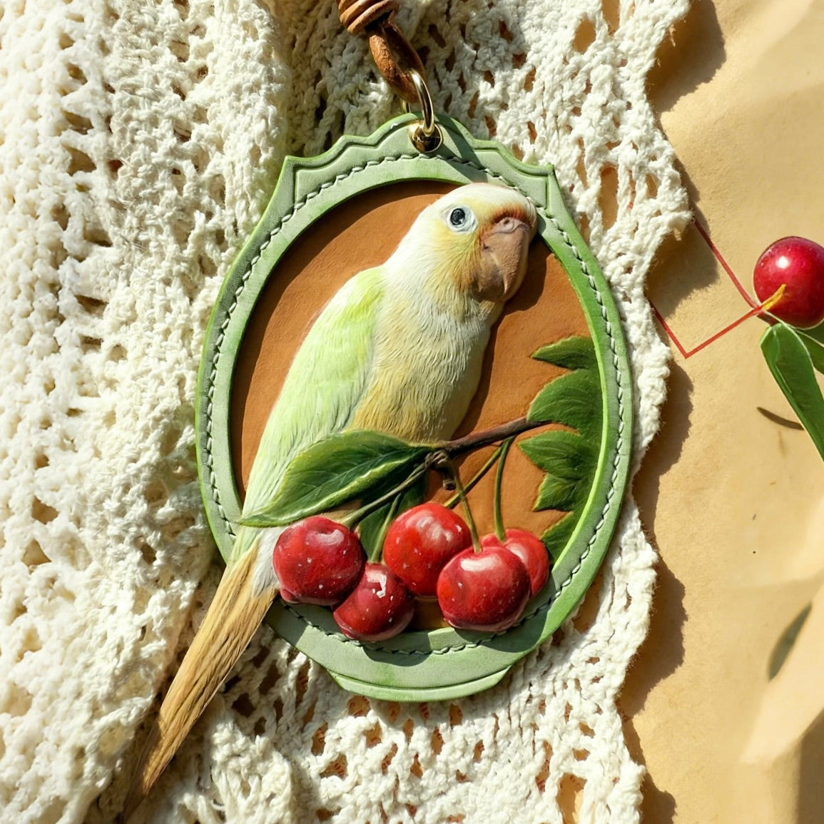 A custom handmade 3D leather-carved pet portrait keychain featuring a realistic light green parrot perched on a branch with red cherries, enclosed in an oval green stitched leather frame on a white lace background.