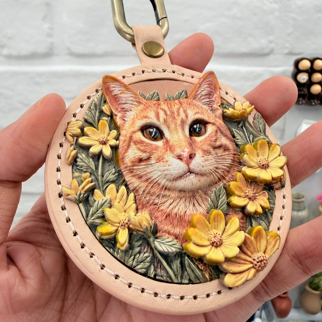 A hand holds a custom round leather keychain featuring a hand-painted 3D relief portrait of an orange tabby cat with yellow flowers, by Memopaw.