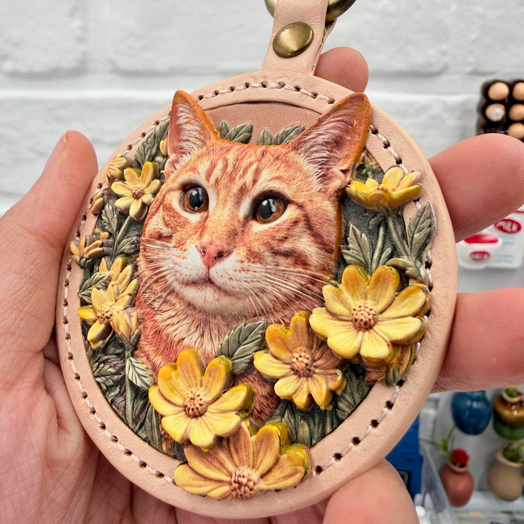 A hand holds a custom round leather keychain featuring a hand-painted 3D relief portrait of an orange tabby cat with yellow flowers, by Memopaw.