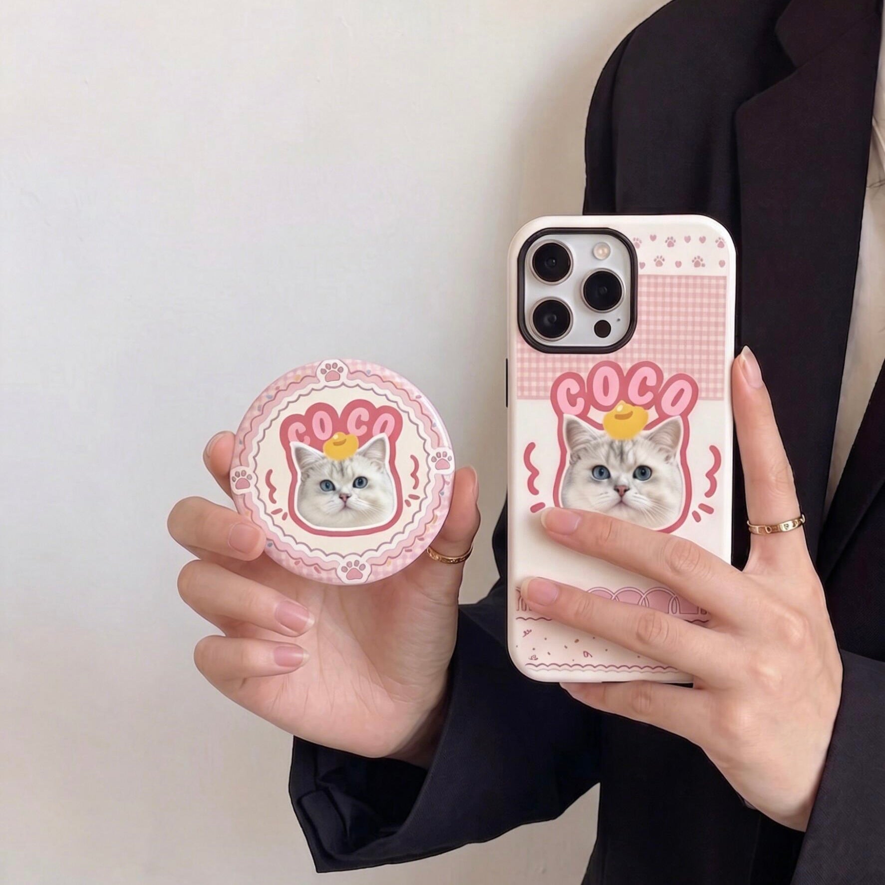 A close-up of a person holding a custom cat photo phone case and a matching round badge, featuring a cute white cat illustration with the name COCO on a pink checkered background.