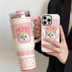 A close-up of a person holding a custom-designed travel tumbler and matching phone case, featuring a personalized cat illustration with the name COCO on a pink checkered background.