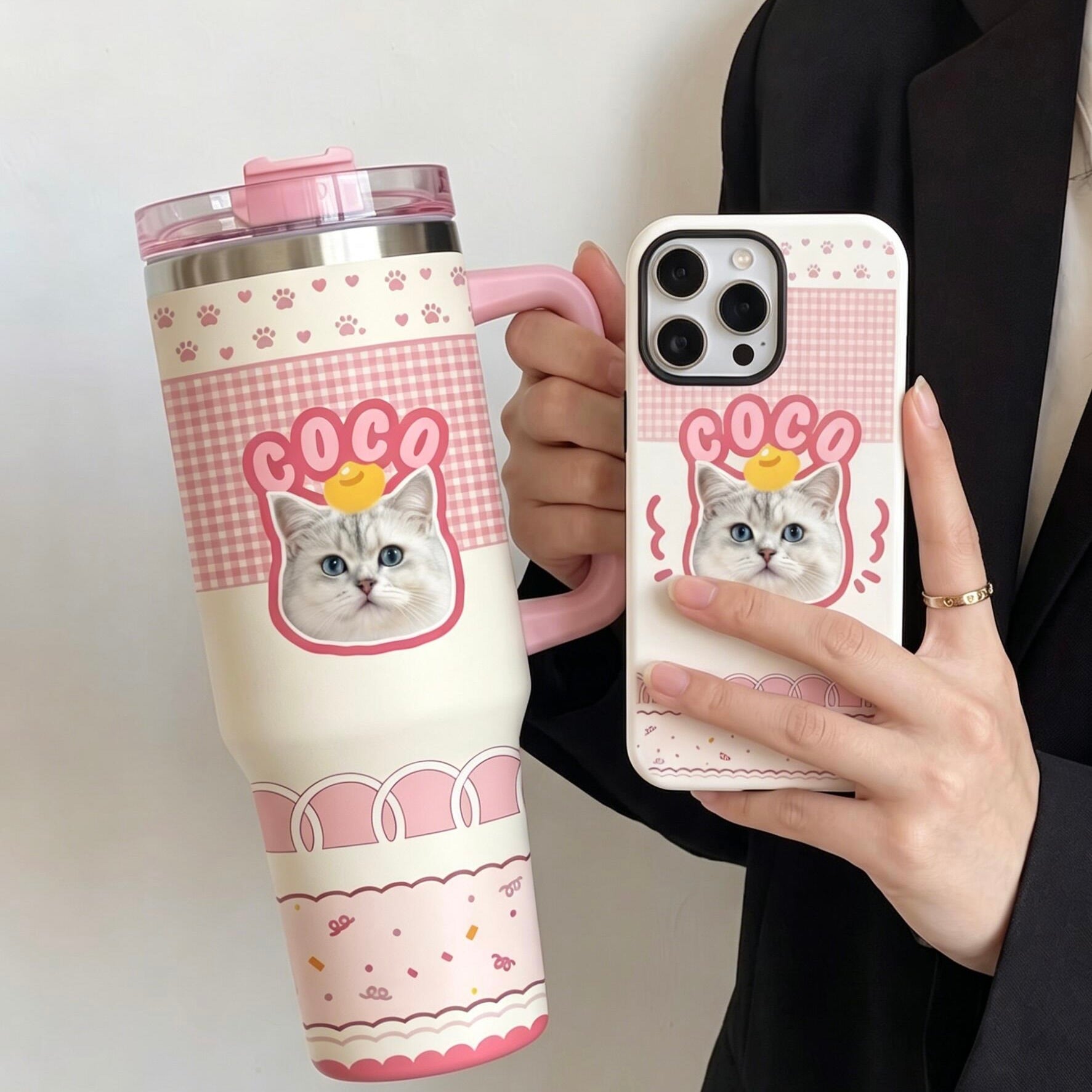 A close-up of a person holding a custom-designed travel tumbler and matching phone case, featuring a personalized cat illustration with the name COCO on a pink checkered background.