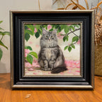 Custom hand-painted oil painting portrait of a fluffy grey cat seated in a dark wooden frame with ornate silver trim, set against a background of green leaves and pink flowers.