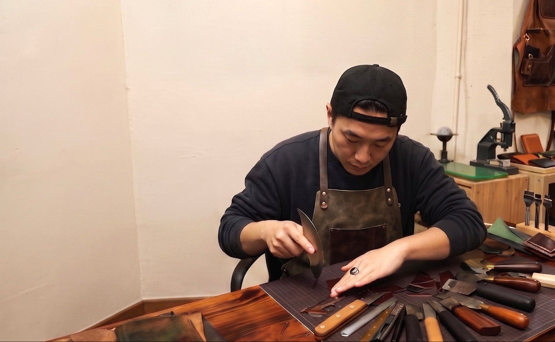 A male leather craftsman in an apron carving raw leather on a workshop desk filled with tools, demonstrating the process of a custom hand-carved leather pet portrait.